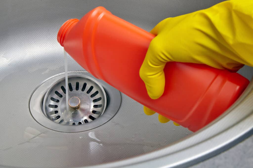 A hand in a yellow rubber glove pours sewer pipe cleaner down the kitchen sink drain.